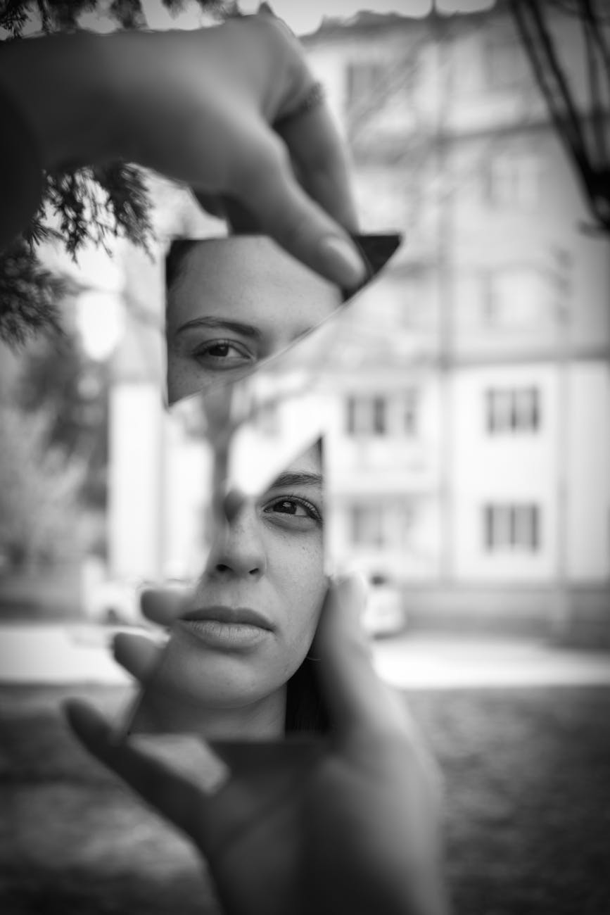 reflection of a woman in small pieces of a broken mirror held by two hands in front of an apartment building in black and white